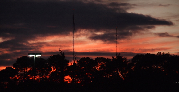 [Behind the silhouette of the pine trees and the skinny cell towers are dramatic orange and dark grey clouds. The clouds between the trees are nearly all orange thus silhouetting the trees.]
